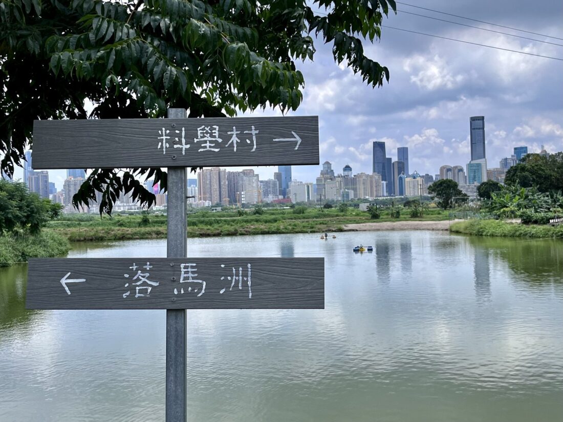Photo of a lake in Hong Kong's New Territories with a sign-post leading to nearby villages in the foreground and the Shenzen skyline in the background.